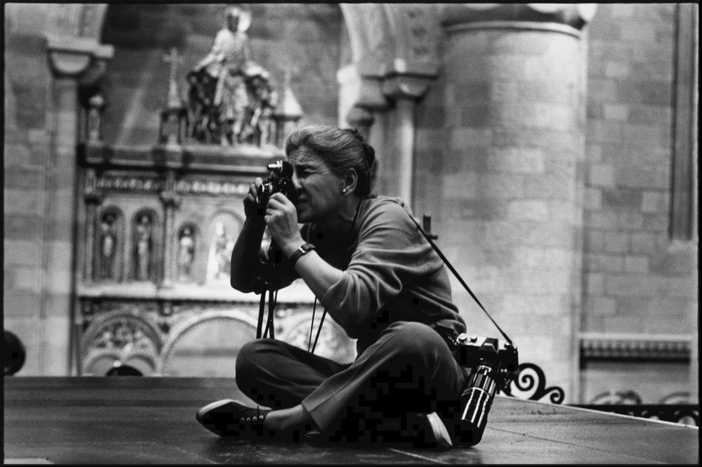 Profile photo of a woman seated cross-legged on the floor of a movie set depicting a medieval castle, peering through a camera viewfinder.