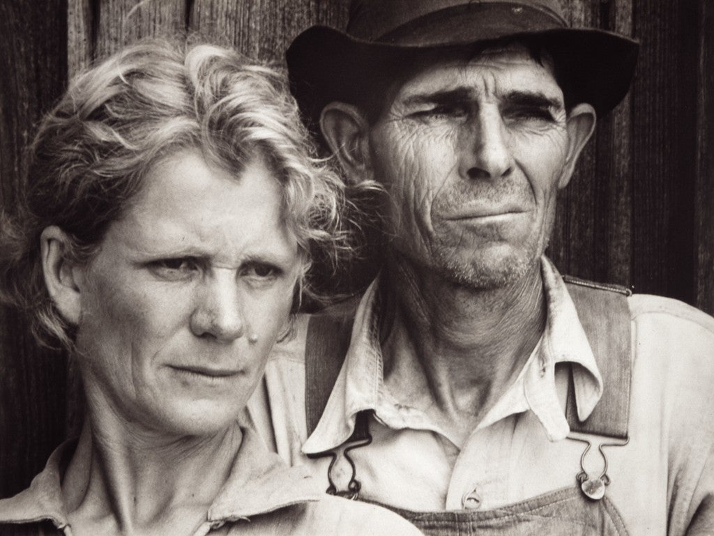 Black and white 1930s portrait of a rural couple standing close together against a wooden wall, their faces lined with strain as they look past the camera.