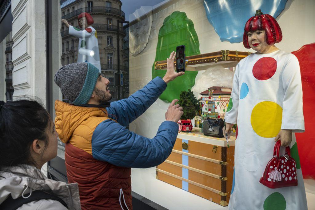A man in a colorful jacket and knit hat takes a photo with his phone of a shop window display featuring a mannequin dressed in a white outfit with large colored dots, holding a red handbag. A woman stands beside him, watching.