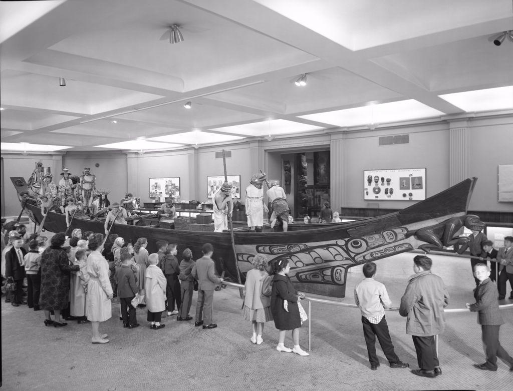 A black-and-white photograph depicting a group of children and adults gathered around North West Coast Canoe displayed in a museum setting. The canoe features detailed artwork with traditional motifs, and several lifelike mannequins dressed in ceremonial attire are positioned in and around the canoe, recreating a historical or cultural scene. The museum walls display additional artifacts and exhibits, creating an educational environment.