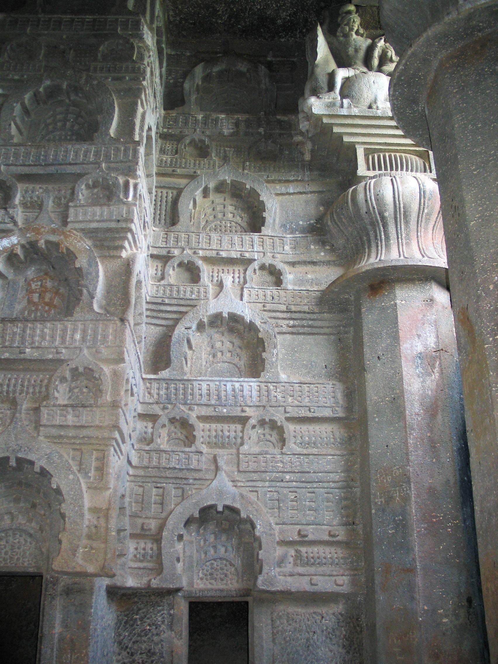 Intricate rock-cut carvings from the Bedsa Caves in Maharashtra, India, dating to the 1st century CE. The wall features a series of ornate chaitya windows, sculpted in repeating patterns, showcasing ancient architectural detailing. A decorative column with a lotus design and a partially visible sculpture of a seated figure are present in the upper corner, highlighting the artistic finesse of the era.