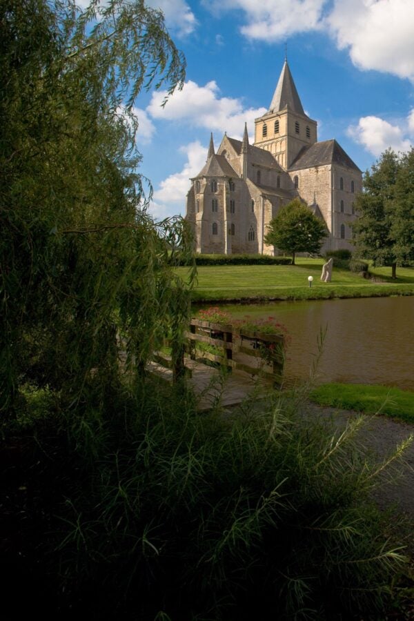 View of a Romanesque abbey church featuring a chevet with rounded apses and a prominent crossing tower, dating to approximately 1032-1072. The stone structure is surrounded by lush greenery and a tranquil pond, with a wooden bridge adorned with flowers in the foreground. The scene is set against a backdrop of a bright blue sky with scattered clouds, emphasizing the historic and serene setting.
