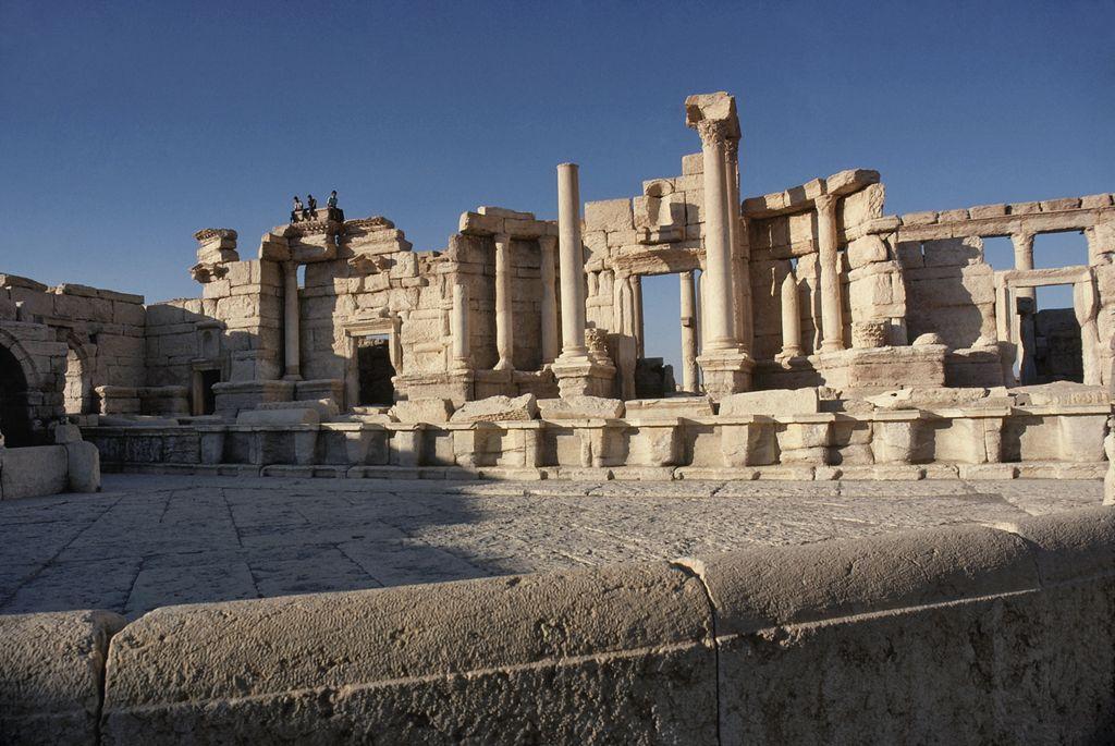 Exterior view of the ancient Roman theatre in Palmyra, Syria, seen from the south. The structure features columns and partially intact stone walls, showcasing classical Roman architectural elements. The ruins sit under a clear blue sky, with figures visible atop the remains, emphasizing the grandeur and historical significance of the site.