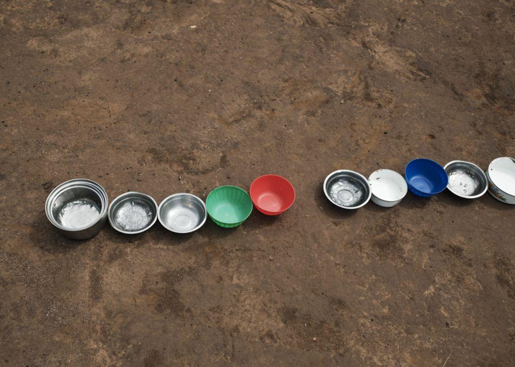 Several colorful plastic bowls and cups arranged in a neat row on a dusty ground, symbolizing scarcity and daily life in a refugee camp.
(Photograph by Salih Basheer, Magnum Photos.)