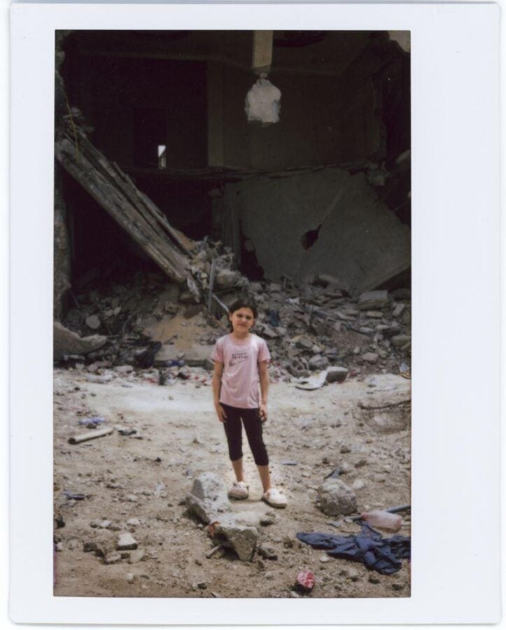 A young girl stands solemnly in front of a bombed and collapsed concrete building in the Jenin Refugee Camp, surrounded by rubble.