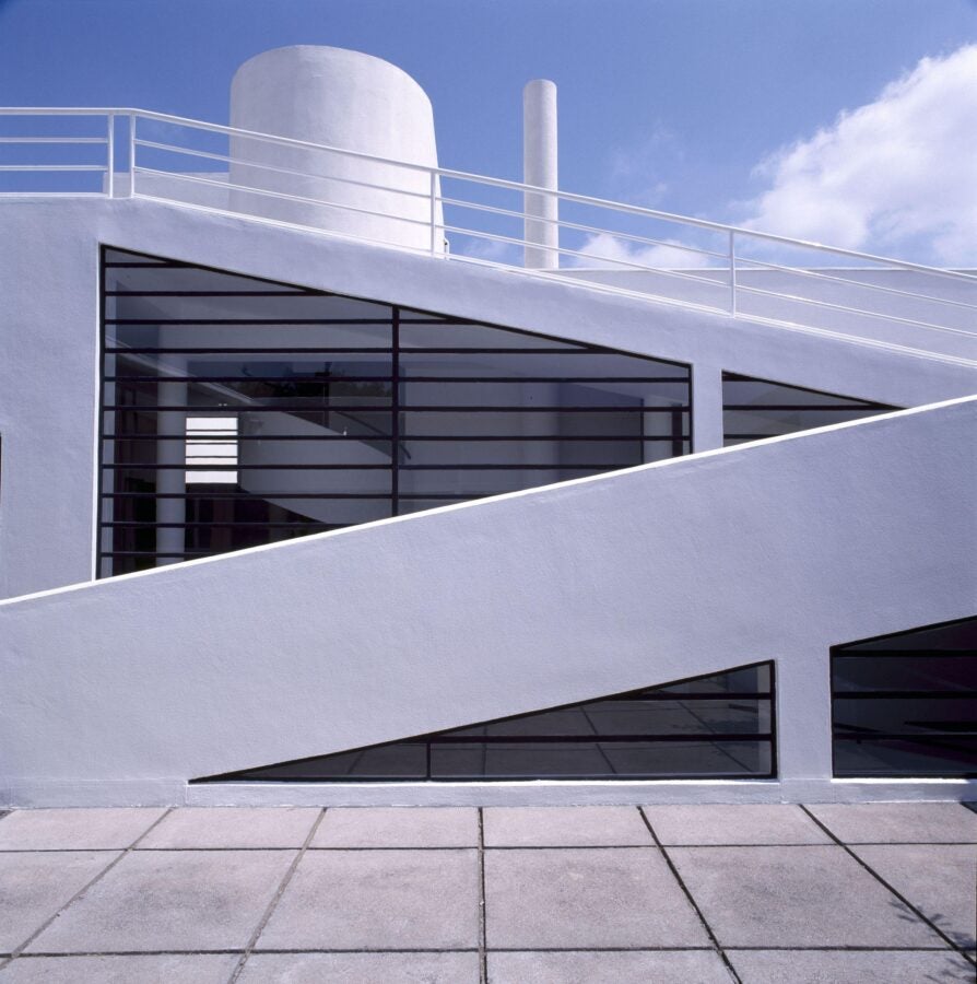 An exterior view of Le Corbusier’s modernist Villa Savoye, showing white geometric walls, angular windows, and a rooftop terrace under a clear blue sky.
(Photograph by Jean-Christophe Ballot, Centre des monuments nationaux.)