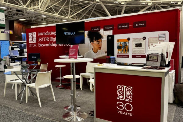 A trade show booth for JSTOR’s Digital Stewardship Service, featuring red branding and a counter with a ‘30 Years’ logo. Behind the counter, a large backdrop displays visuals related to metadata creation and collection management. The booth includes a high table with stools, a glass table with chairs, two monitors showing the ITHAKA logo, and promotional materials neatly arranged on the counter.