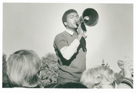 A black-and-white photograph of a man speaking passionately through a megaphone at an outdoor gathering. He wears a light shirt under a dark sweater and stands above a crowd, with trees visible in the background. Several people in the foreground, some with light-colored hair and glasses, listen closely and raise their hands.