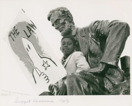 A young Black boy sits beside a large bronze statue of Abraham Lincoln, holding a protest sign with a drawing of a Ku Klux Klan hooded figure labeled “THE LAW.” The handwritten caption at the bottom reads “Support Alabama 1963.”