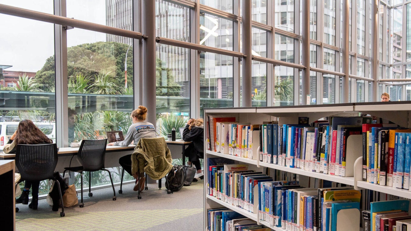 Students study at desks beside large windows overlooking greenery inside the library at Victoria University of Wellington.