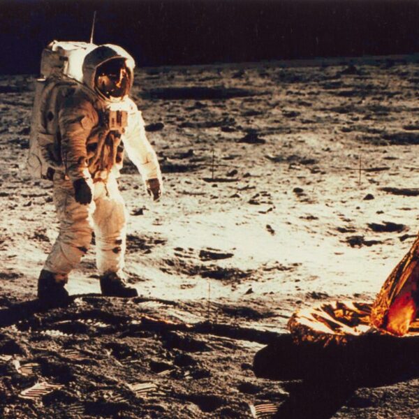Astronaut in a white spacesuit standing on the moon’s surface near a lander footpad, with footprints and lunar soil illuminated against the dark sky.
