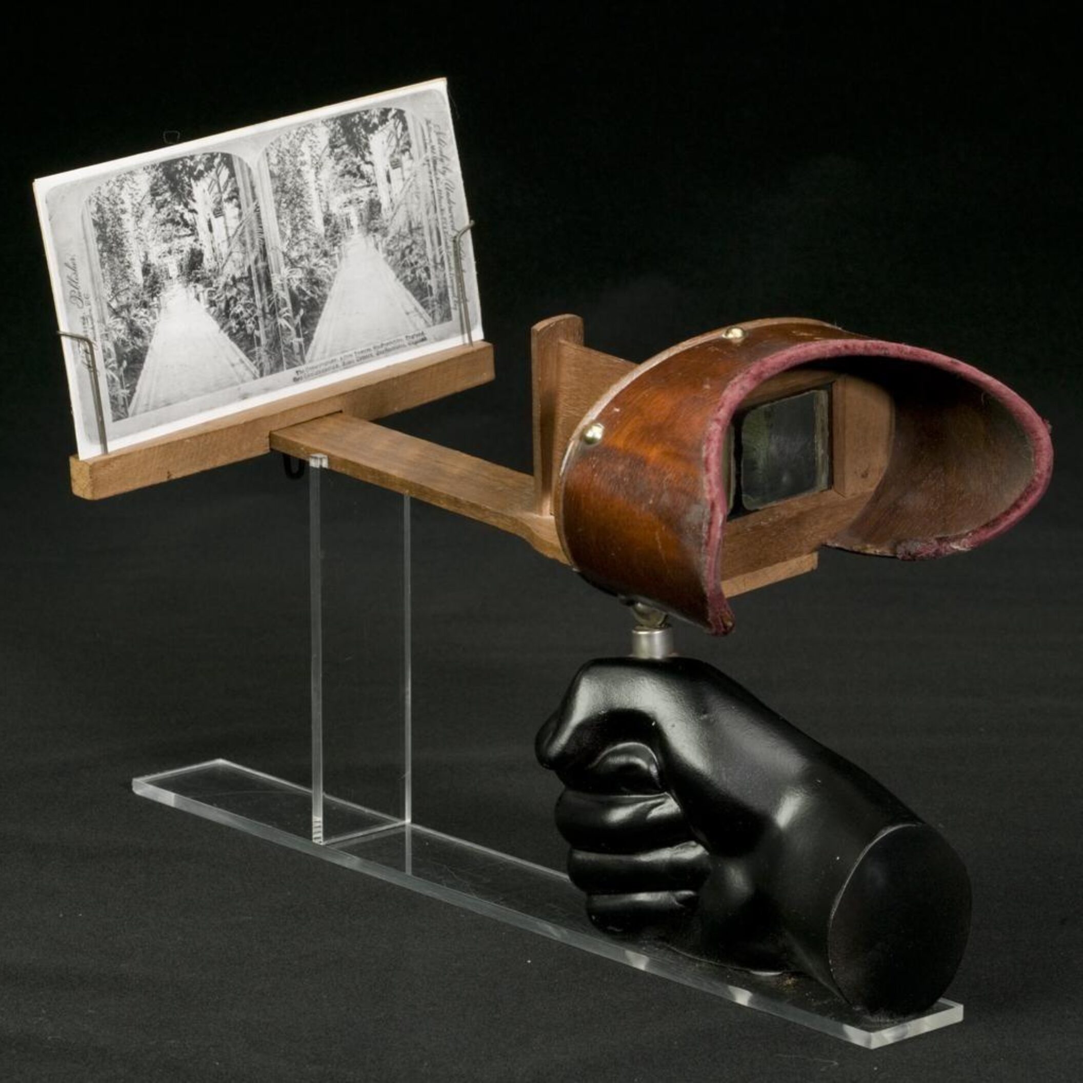 Handheld stereoscope viewer with wooden frame and leather hood, holding a black-and-white stereoscopic photograph.