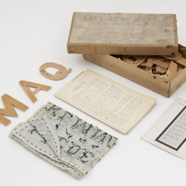 A vintage educational kit for teaching Braille, including a worn cardboard box filled with wooden alphabet letters, several loose wooden letters with Braille markings, typed instruction sheets, a stitched fabric sampler, and a printed Braille chart arranged on a white surface.