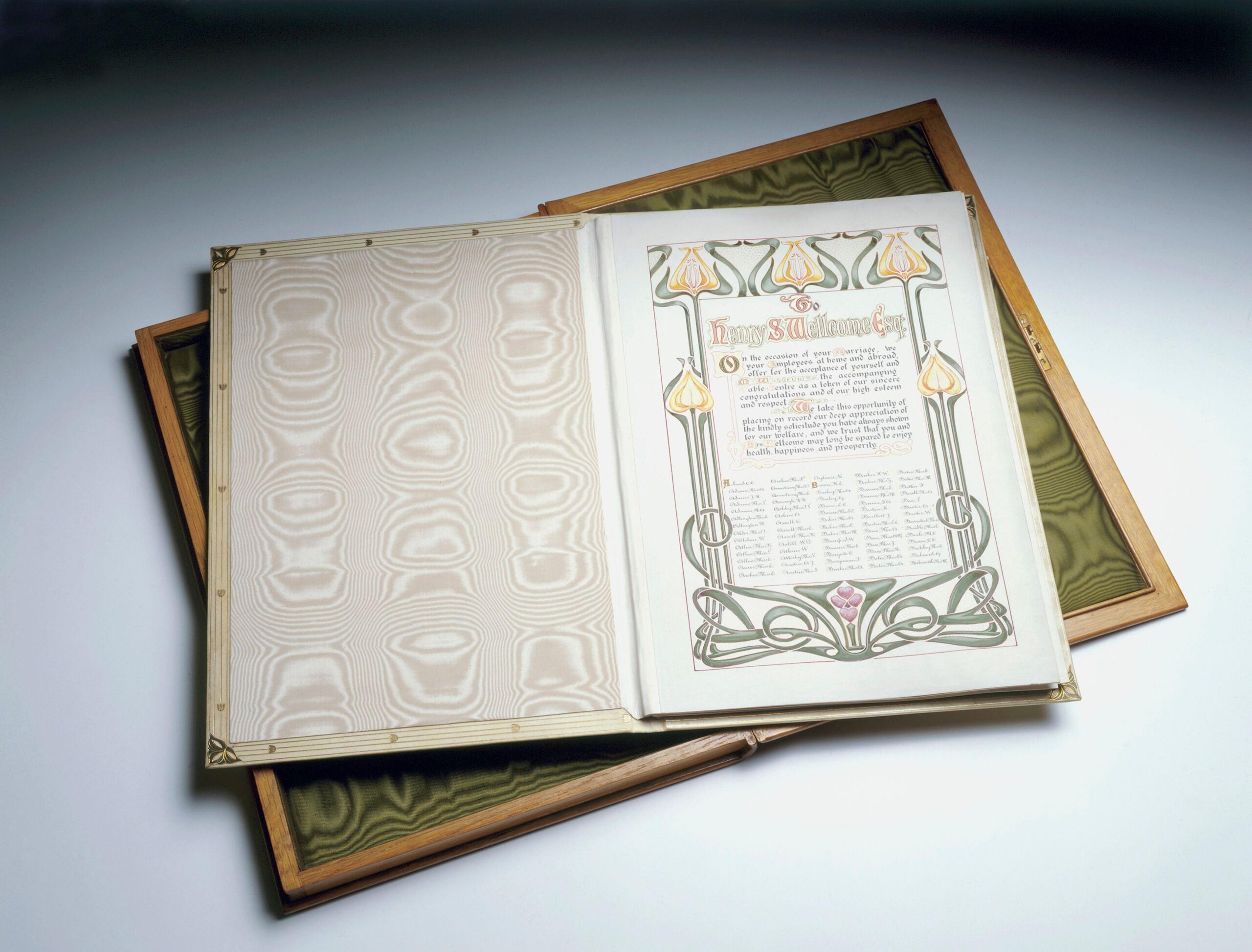 An open illuminated manuscript with Art Nouveau–style borders and floral motifs rests on top of another decorative book. The visible page features elegant calligraphy framed by stylized flowers and leaves in green, gold, and pink tones. The book’s cover and the background surface are shades of beige and green, and the scene is softly lit to emphasize the intricate design and texture of the paper.