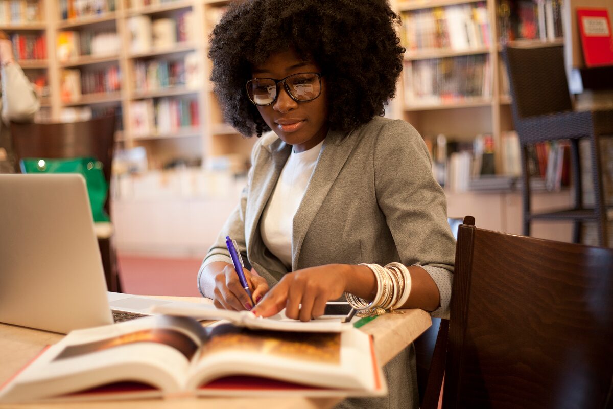 Person writing notes while studying at a library table with open books and a laptop.
