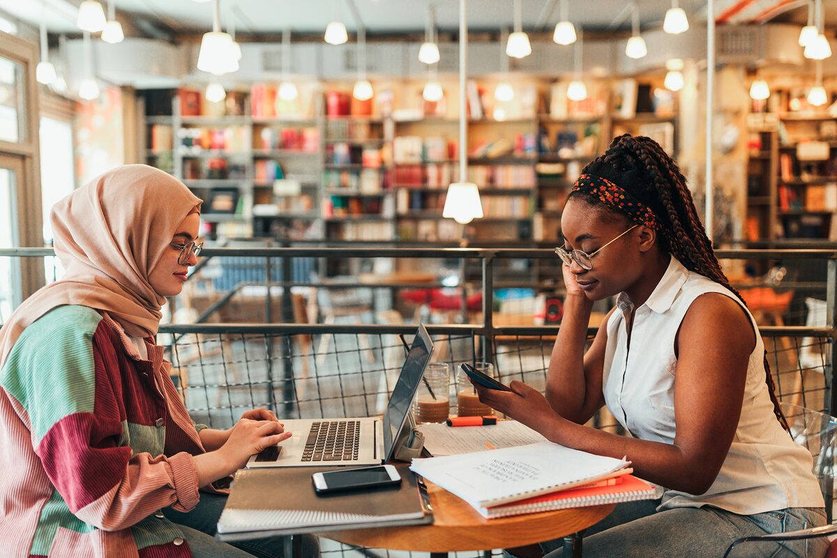 Two students sit in a cozy café surrounded by books, studying together with laptops and notebooks.