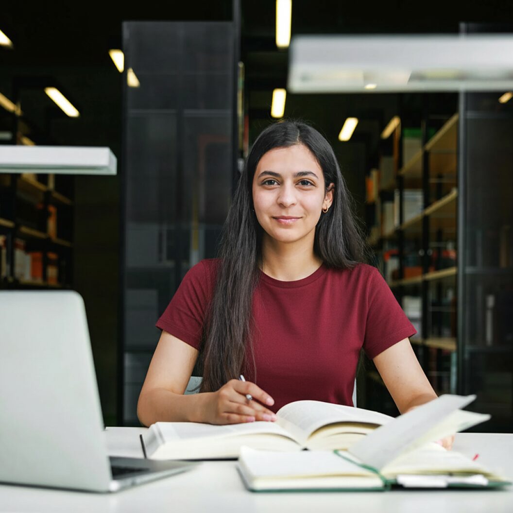 Smiling student seated at a library table with open books and a laptop, representing research and study on the JSTOR platform.