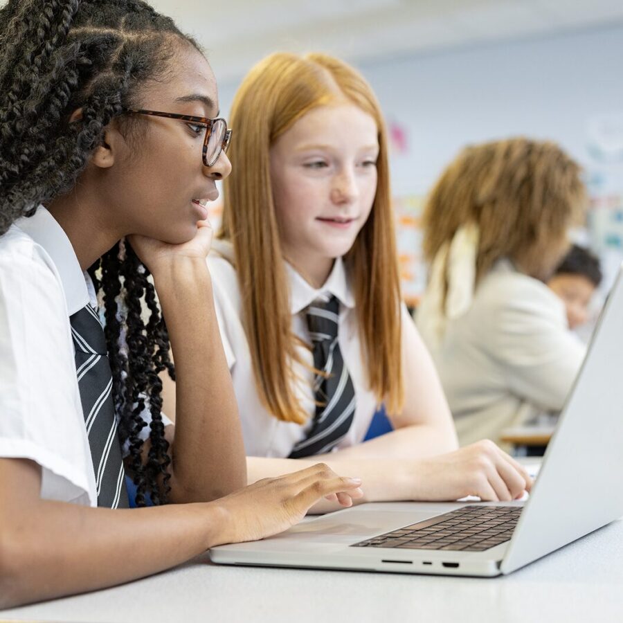 Two secondary school students in uniforms collaborating on a laptop in a classroom setting, focused on their work and discussing research.