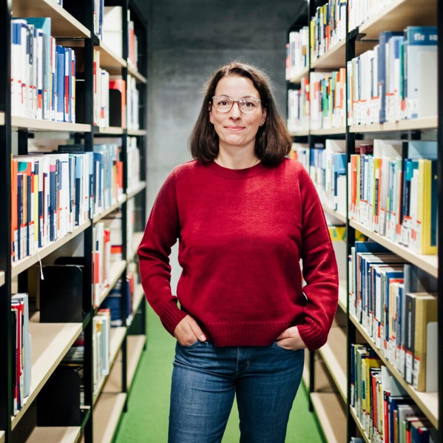 A person wearing a red sweater stands confidently between two rows of bookshelves in a modern library.