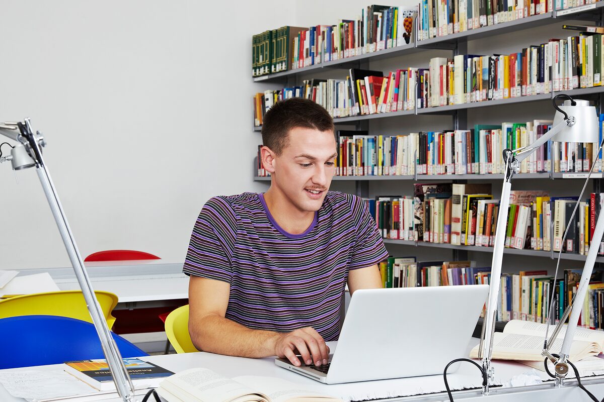 A student sits at a table in a library, working on a laptop surrounded by books and study lamps.