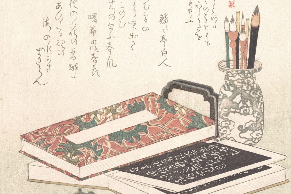 Japanese woodblock print showing a decorative brush pot filled with calligraphy brushes beside stacked patterned books and writing tools. Delicate flowers and handwritten Japanese text appear around the still-life arrangement.