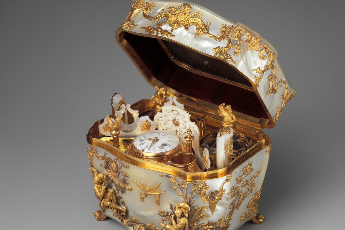 An ornate 18th-century nécessaire box with its lid open, featuring mother-of-pearl panels and elaborate gilded decoration. Inside are finely crafted grooming tools and a small clock, all set within a luxurious gold-trimmed interior.