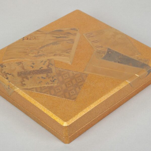 A gold Japanese writing box with a flat rectangular lid decorated in lacquer with layered illustrated books.