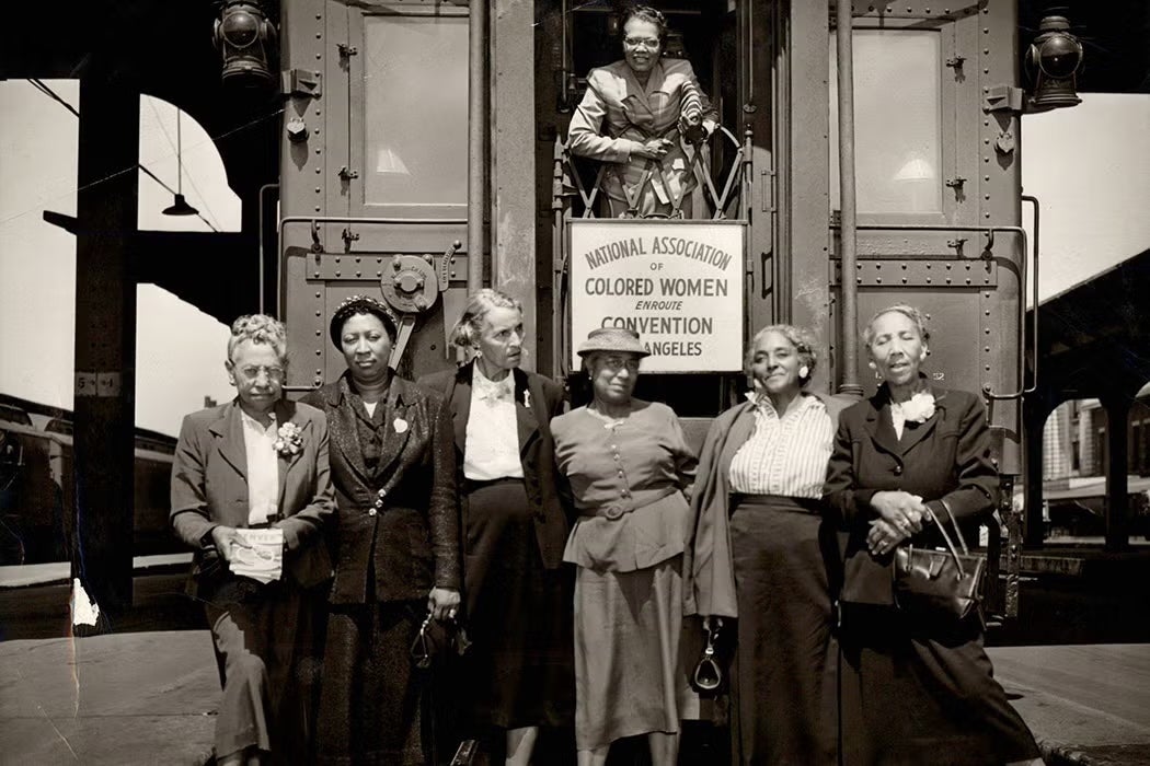 Group of Black women standing beside a train car under a sign reading “National Association of Colored Women en route to Convention, Los Angeles,” with one woman looking out from the train doorway.