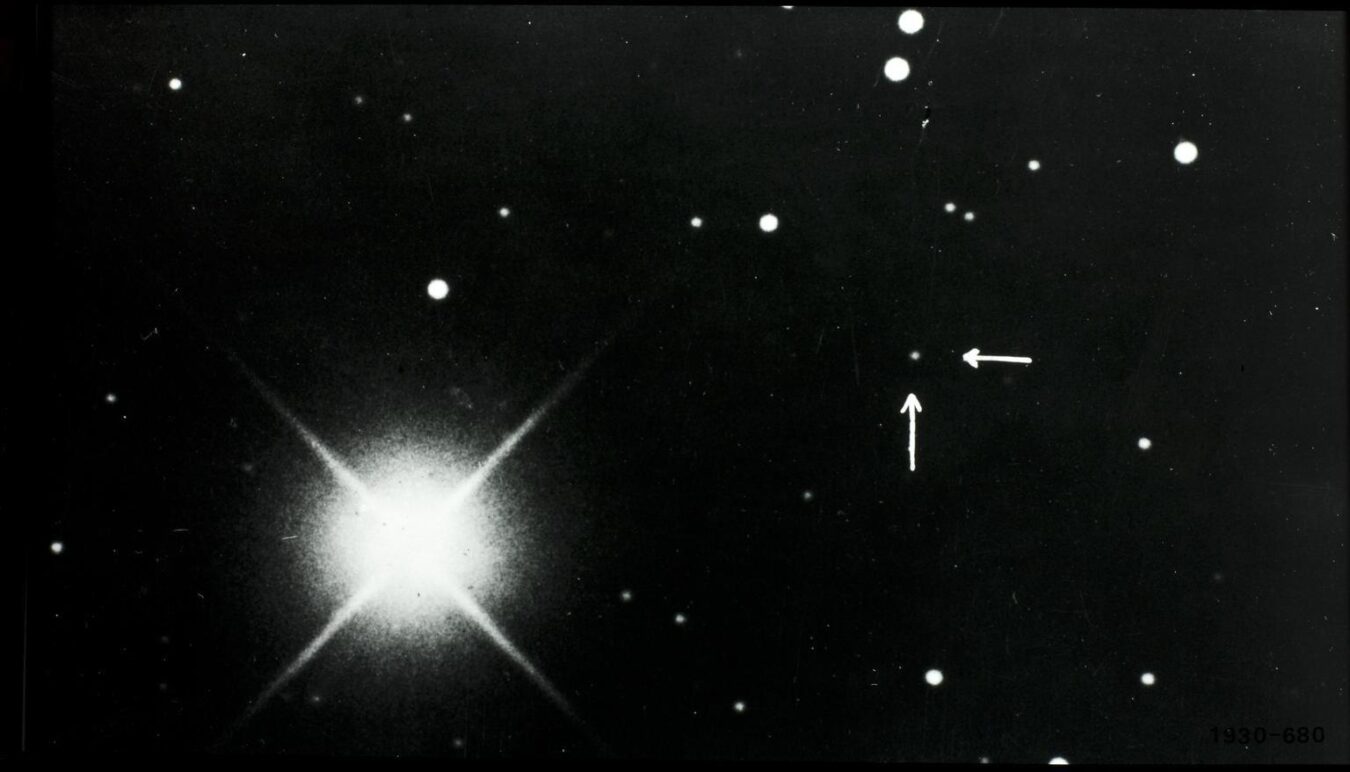 Black-and-white astronomical photograph showing a bright star with diffraction spikes and surrounding points of light; arrows indicate a faint object identified as Pluto.