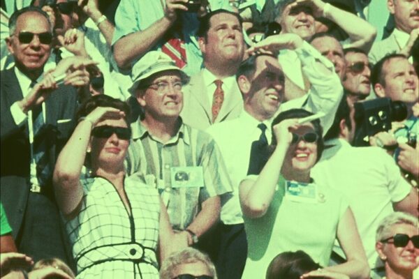 Crowd of spectators at the Apollo 11 launch site in 1969, many shading their eyes or wearing sunglasses as they look upward; several hold cameras while watching the rocket lift off.