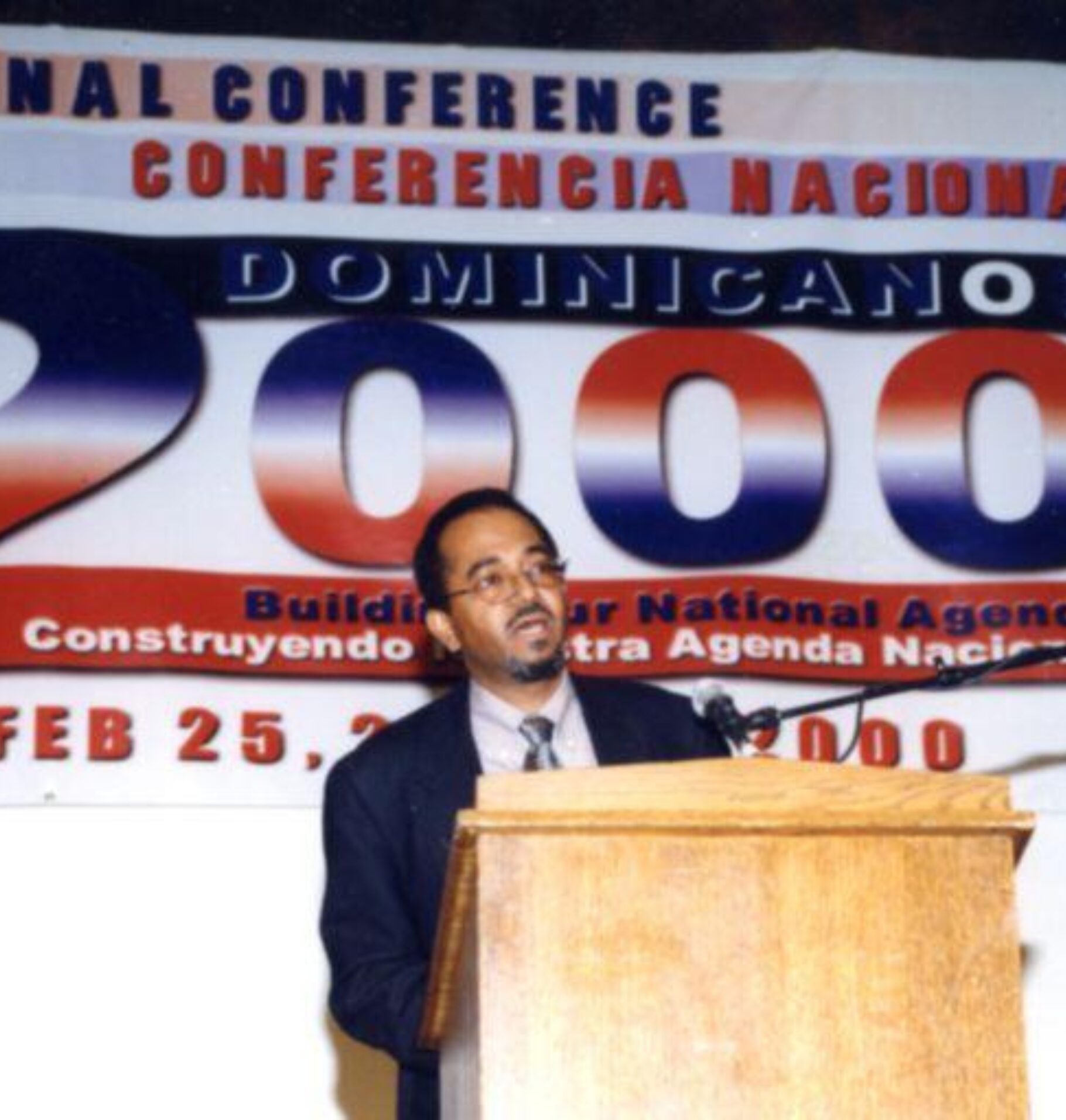 Man speaking at a podium beneath a large banner reading “National Conference Dominicanos 2000: Building Our National Agenda,” dated February 25, 2000.