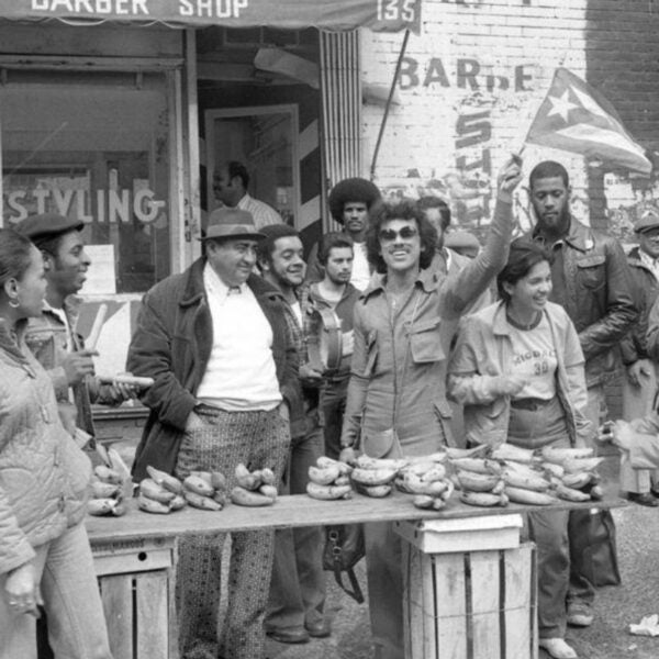 Black-and-white photo of a group of people gathered around a table outdoors, one person holding a Puerto Rican flag, in front of a neighborhood barbershop during a 1971 student strike.
