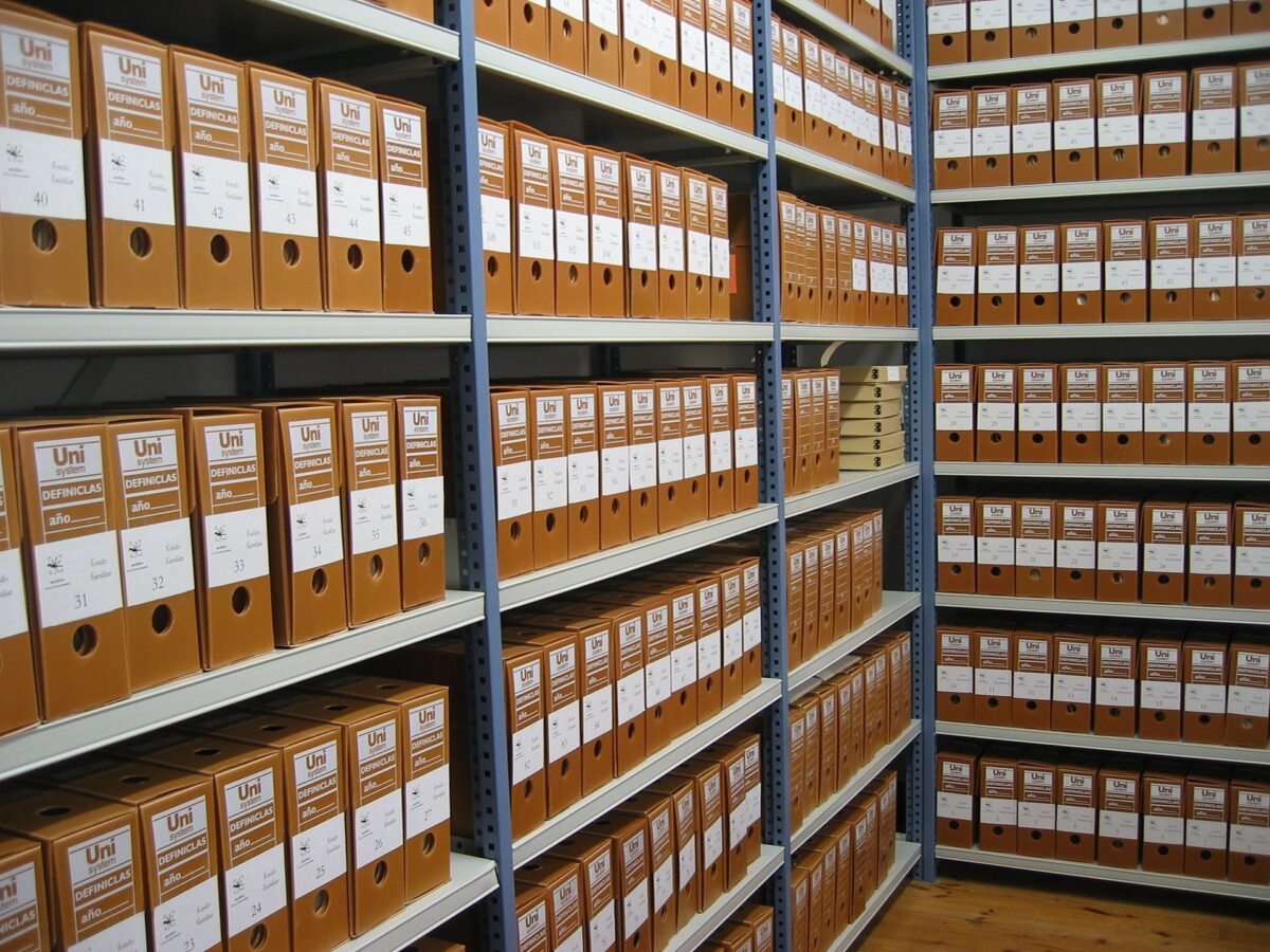 Rows of orange archival storage boxes with white labels neatly arranged on gray metal shelving in a records storage room. Each box is labeled and numbered, and the shelves extend along a wooden floor aisle.