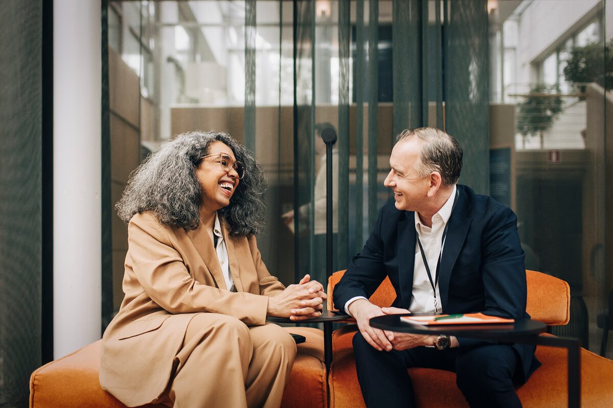 Two colleagues in business attire sit in a modern office lounge, smiling and engaged in a friendly conversation across a small table.