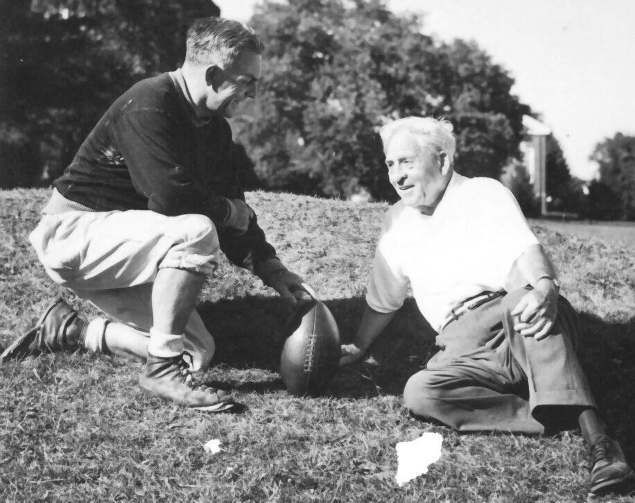 Black-and-white photograph of two men outdoors on a grassy field. One man in vintage football attire kneels beside a football while speaking with an older man seated on the ground. Trees and campus-like buildings appear in the background.