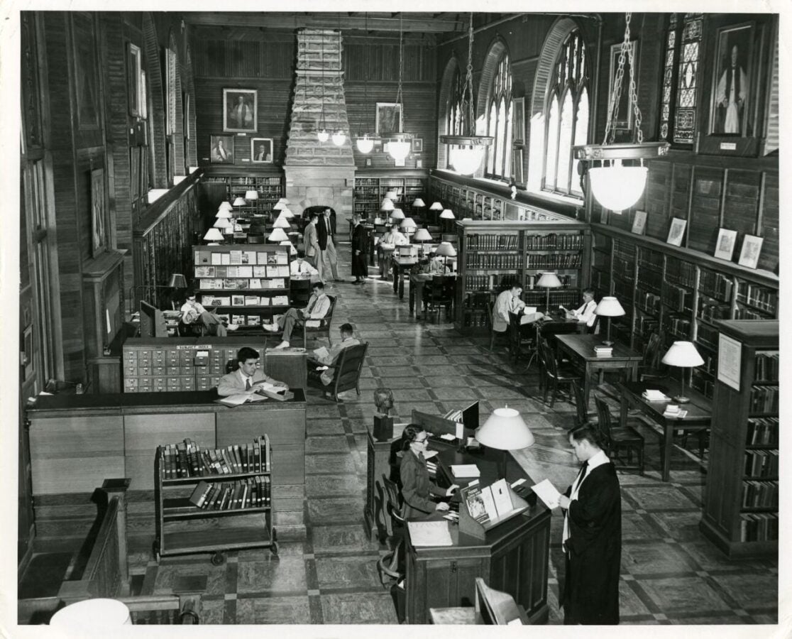 Black-and-white photograph of a large collegiate library reading room with tall arched windows, bookshelves, study tables, hanging light fixtures, and a central stone fireplace. Students and staff are reading, studying, and working throughout the space.